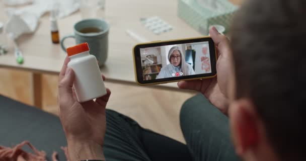 여성 성숙 한 의사와 이야기하고 비디오 폰 앱을 사용하는 사람의 어깨 너머로 보기. 온라인상 상담을 받거나 소파에 앉아 있는 동안 플라스틱 알약 병을 들고 있는 사람은. — 비디오
