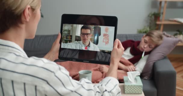 Overschouderzicht van een vrouw die een tablet vasthoudt en in gesprek is met een mannelijke arts. Mam houdt online medisch consult terwijl zieke zoon de temperatuur meet en op de bank ligt. Concept telegezondheid. — Stockvideo