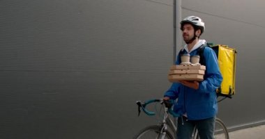 Siyah duvarın yanında yürürken sırt çantasıyla bisikleti iten erkek teslimatçının ön görüntüsü. Pizza kutuları ve kahve fincanları taşıyan koruyucu kasklı adam..