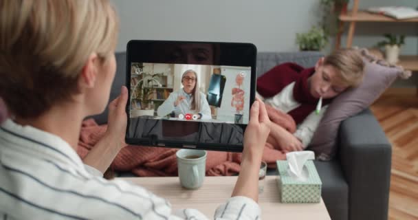 Rückansicht einer Frau, die ein Tablet in der Hand hält und sich online ärztlich beraten lässt, während ihr kranker Sohn zu Hause auf dem Sofa liegt. Konzept von Krankheit und Telemedizin. — Stockvideo