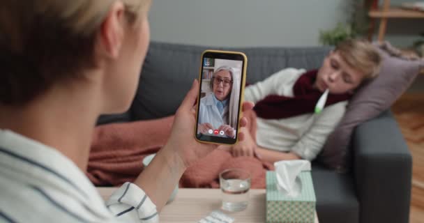 Matka drží smartphone a mluví se starší doktorkou. Žena má online lékařskou konzultaci, zatímco její syn leží na pohovce s teploměrem. Koncepce telemedicíny. — Stock video