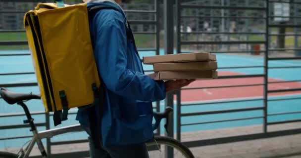 Vue latérale du livreur avec sac à dos de transport poussant le vélo tout en marchant près du terrain de sport. Courrier portant des boîtes à pizza tout en livrant la commande au client. — Video