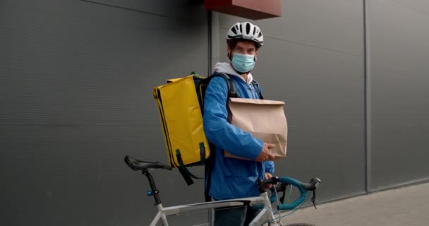 Cyklistický kurýr v lékařské masce a cyklistické helmě zvedá hlavu a dívá se do kamery. Guy s transportní taškou a papírovým balíčkem drží kolo, zatímco stojí na ulici. — Stock video