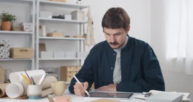 Tabletinde stilus olan bıyıklı genç bir erkek grafik tasarımcısının portresi. 30 'lu yaşlarda bir adam sıradan kıyafetler giyip masa başında çalışırken kameraya bakıyor..