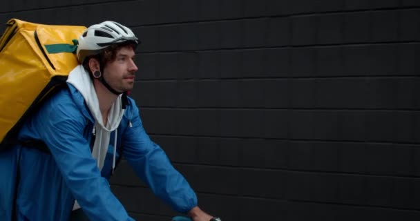 Человек-курьер с сумкой для транспортировки еды на плече на велосипеде на улице. Парень в защитном велосипедном шлеме доставляет заказ клиенту. Концепция доставки продуктов питания. — стоковое видео