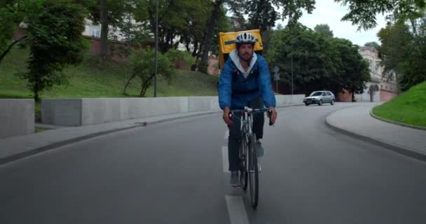 Mužský kurýr v ochranné přilbě s izolační taškou na přepravu potravin jízdní kolo na silnici.Chlap s bezdrátovými sluchátky doručující objednávku zákazníkovi na kole. Auto na pozadí. — Stock video