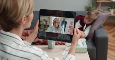 Tablet tutan ve olgun kadın doktorla iletişim kuran kadının omuz üstü görüntüsü. Annesi hasta oğlu kanepede yatarken internetten tıbbi konsültasyon alıyor. Tele-sağlık kavramı.