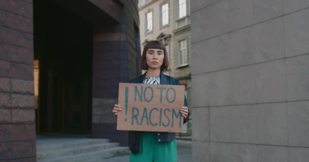 Egy fiatal nő, aki nemet mond a rasszista kifejezésmódú kartonpapírra, miközben a város utcájában áll. Vonzó hipszter lány támogatja a rasszizmus elleni kampányt. Az egyenlő emberi jogok fogalma. Közelíts rá!. — Stock videók