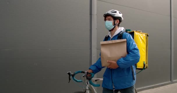 Vista de colheita da pessoa de entrega em máscara médica com mochila de transporte de alimentos empurrando bicicleta na rua. Cara no capacete protetor transportando pacote de papel enquanto caminhava perto da parede preta. — Vídeo de Stock