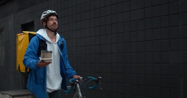 Coursier masculin joyeux en casque de protection avec sac de transport alimentaire transportant du café et poussant le vélo dans la rue. Guy souriant et marchant tout en livrant la commande au client. — Video