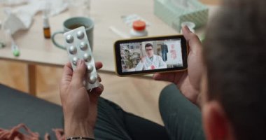Cep telefonuyla hastaya danışmanlık yapan genç bir doktorun omuz hizasında. Elinde hap tutan ve koltukta otururken akıllı telefon kullanan hasta. Teletıp kavramı.