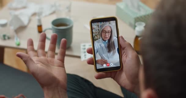 Medico donna matura in occhiali consultando paziente tramite videochiamata su smartphone e scrivendo dettagli. Sopra spalla primo piano visualizzazione dello schermo mobile. Concetto di telemedicina. — Video Stock