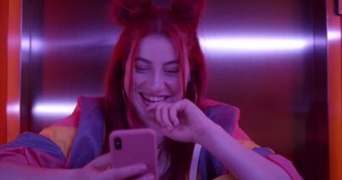 Akıllı telefon kullanan ve neon ışıkta otururken gülümseyen çekici bir kadın. Modaya uygun saç stili olan mutlu bir kadın telefon ekranına ve kameraya bakıyor..