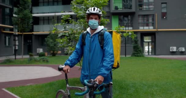 Portrét mužského kurýra v ochranné lékařské masce a cyklistické helmě stojící v rezidenčním komplexu. Guy s přepravou potravin taška drží kolo a hledá kameru venku. — Stock video