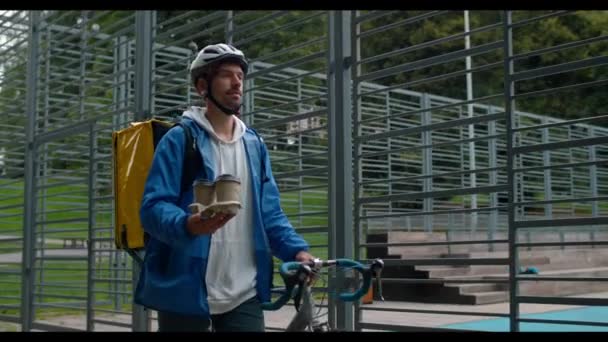 Muž cyklistický kurýr s přepravou potravin batoh tlačí kolo při chůzi v blízkosti sportovního hřiště. Muž v ochranné cyklistické helmě nesoucí šálky na kávu při dodání objednávky. — Stock video