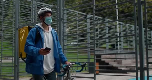 Mensajero masculino en máscara médica con bolsa de transporte empujando bicicleta cerca del campo deportivo. Guy en casco de ciclismo de protección mirando el mapa en el teléfono inteligente mientras entrega el pedido al cliente. — Vídeo de stock