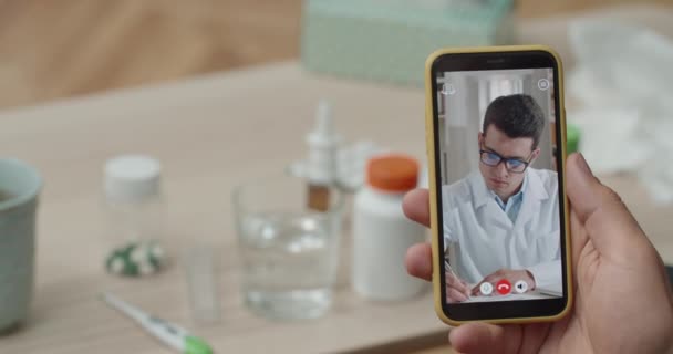 Ung manlig läkare konsultera patienten genom videosamtal på smartphone och skriva detaljer. Över axeln närbild mobiltelefon skärmvy. Begreppet telemedicin. — Stockvideo