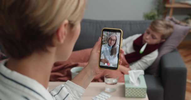 Visão traseira da mulher segurando smartphone e tendo consulta médica on-line enquanto seu filho doente deitado no sofá em casa. Conceito de doença e telessaúde. — Vídeo de Stock