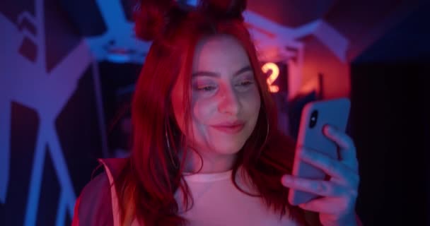 Zamknij widok atrakcyjnej młodej kobiety za pomocą smartfona i patrząc na ekran. Kobieta uśmiechnięta podczas spaceru korytarzem z neonowymi światłami i świecącymi liczbami. — Wideo stockowe