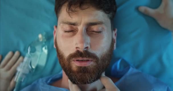 Закрытие рук медиков, надевающих больному бородатому человеку кислородную маску. Верхний вид бессознательного парня 30-х годов бормочущего, лежащего на тележке с медицинским транспортом по дороге в больницу. — стоковое видео