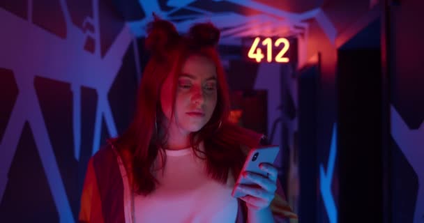 Zamknij widok poważnej kobiety korzystającej z telefonu komórkowego. Przystojny Milenium kobieta z modnej fryzury patrząc na ekran telefonu podczas spaceru w korytarzu z neonów. — Wideo stockowe
