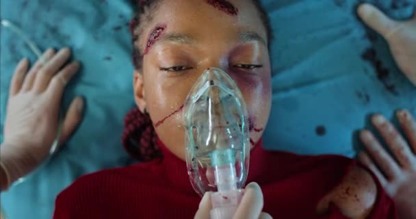 Femme afro-américaine avec blessures et masque à oxygène allongé sur une civière de l'hôpital en ambulance. Vue rapprochée de la jeune fille avec du sang et des blessures sur sa tête ambulanciers transportant à la clinique. — Video