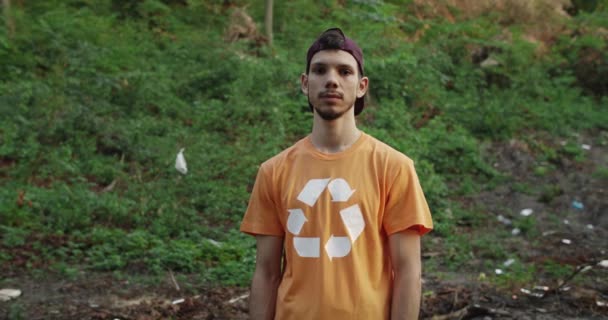 Porträtt av ung kille med mössa på huvudet bär t-shirt med återvinning symbol på det tittar till kameran. Millennial manliga eko aktivist står i skogen full av skräp. Ekologiskt begrepp. — Stockvideo