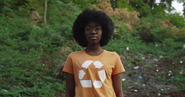 Porträt einer jungen Afroamerikanerin, die ein T-Shirt mit Recycling-Symbol trägt und in die Kamera blickt. Millennial-Öko-Aktivistin mit Brille steht in Park voller Müll. — Stockvideo