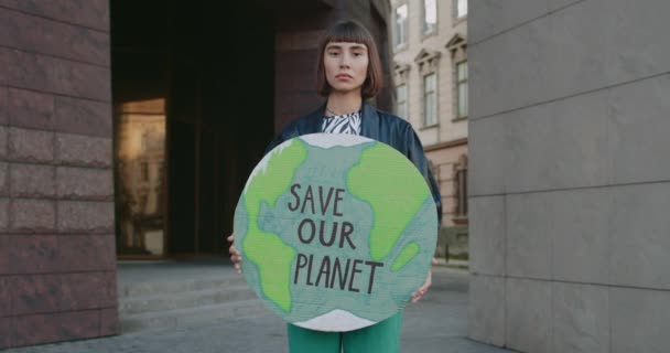 카톤 플래카드를 가진 진지 한 여자가 친환경 운동을 지지하면서 카메라를 쳐다 봅니다. 젊은 여자가 길거리에 서 있는 동안 지구의 글쓰기를 구할 수있는 지구 모델을 들고 있습니다.. — 비디오