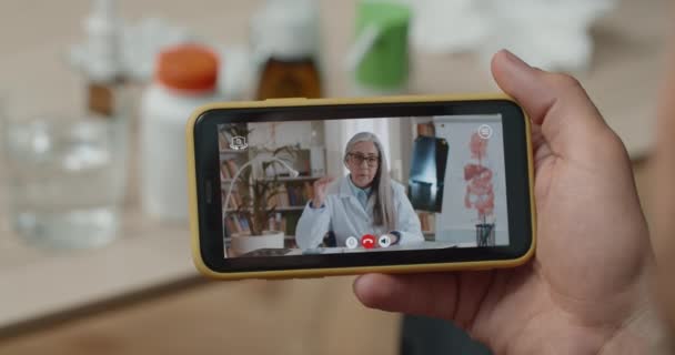 스마트폰을 들고 성숙 한 여성 의사와 이야기하는 사람들의 모습을 어깨 너머로 바라봅니다. 환자가 온라인 진료를 받는 모습을 확대 해서 보 세요. 원격 의료의 개념. — 비디오