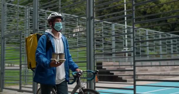 Cara em máscara médica com saco de transporte empurrando bicicleta perto do campo esportivo. Correio de bicicleta em capacete de ciclismo protetor transportando xícaras de café de papel enquanto entrega ordem ao cliente. — Vídeo de Stock