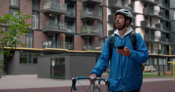 Mensageiro de bicicleta masculino com saco de transporte olhando ao redor enquanto procura endereço de entrega. Cara no capacete de proteção usando smartphone e empurrando bicicleta no complexo residencial. — Vídeo de Stock