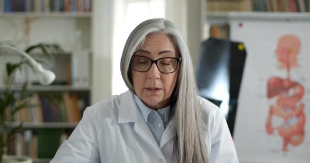 Crop view of female doctor talking and gesturing while having online consultation. Mature woman in glasses and white gown communicating with patient and looking to camera. — Stock Video