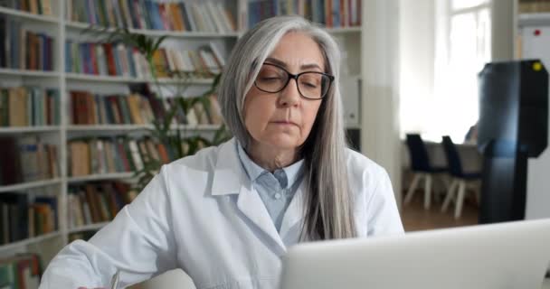 Close-up van geconcentreerde dame in bril en witte jurk typen en invullen van medische documentatie. Oudere vrouwelijke arts werkt met laptop op kantoor. Concept van routine en werk. — Stockvideo