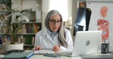 İnternette danışmanlık yaparken konuşan ve dizüstü bilgisayara bakan yaşlı bir kadın. Video görüşmesi yaparken ve ofiste otururken hastayla iletişim kuran olgun kadın doktor..