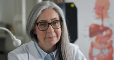 Gülümseyen bir kadının portresi başınızı çevirir ve tıbbi ofiste otururken kameraya bakar. Gözlüklü kadın doktor ve beyaz profesyonel soyguna yakından bakın. Vesikalık fotoğraf kavramı.