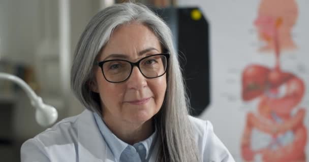 Portrait of smiling woman turning head and looking to camera while sitting in medical office. Close up view of female doctor in glasses and white professional rob. Concept of headshot. — Stock Video