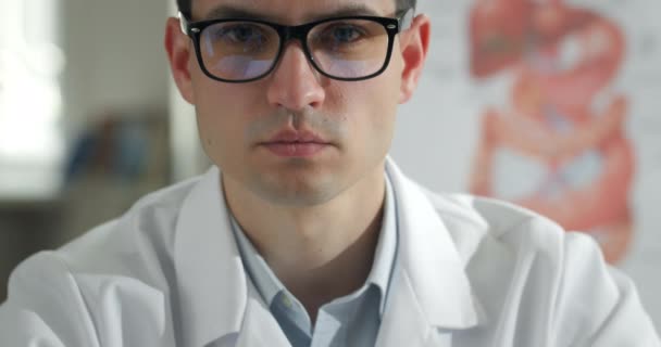 Close up view of concentrated male doctor looking to camera in medical office. Portrait of serious young man in white professioanal coat and glasses. — Stock Video