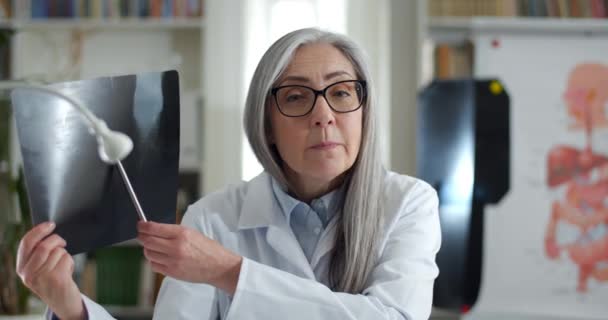 안경과 흰색 강도질을 한 전문직 여성이 온라인 통화를 하면서 엑스레이 사진을 보여 주고 이야기하는 모습이 눈에 띈다. 여자 의사가 카메라를 찾고 있어. 전신 건강에 대한 개념. — 비디오