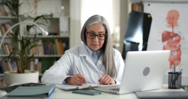 Çevrimiçi danışmanlık yaparken beyaz önlüklü neşeli yaşlı kadın tıbbi form dolduruyor. Gözlüklü kadın doktor görüntülü konuşma yaparken konuşuyor ve bilgi yazıyor.