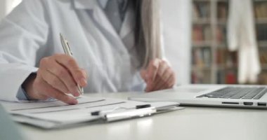 Beyaz profesyonel önlüklü bir kadının tıbbi belgeleri yazıp doldurması. Masada otururken dizüstü bilgisayarla çalışan kadın doktor. Rutin ve çalışma kavramı.