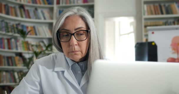 Close-up zicht op oudere vrouw in bril en witte rob typen en kijken naar laptop scherm.Volwassen vrouwelijke arts werken met personal computer in het medisch kantoor. Begrip routine. — Stockvideo