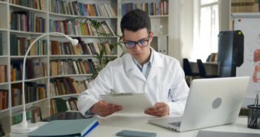 Erkek doktor ofiste otururken tıbbi dokümanları okuyor ve başını sallıyor. Gözlüklü genç bir adam ve dizüstü bilgisayarında yazan beyaz profesyonel bir elbise. Tıp çalışanı kavramı.