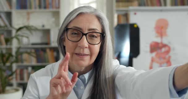 Close up of elderly female doctor in glasses waving and talking while holding smartphone. Mature woman in white gown having online consultation in medical office. Concept of telemedicine. — Stock Video