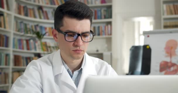 Primo piano di uomo concentrato in occhiali e cappotto bianco professionale digitando e guardando lo schermo. Giovane medico di sesso maschile che lavora con il computer portatile in carica. Concetto di routine del lavoratore medico. — Video Stock