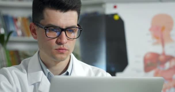 Close up of male doctor in glasses looking at laptop screen while working. Man in white professional rob while sitting in medical office. Concept telemedicine and telehealth. — Stock Video