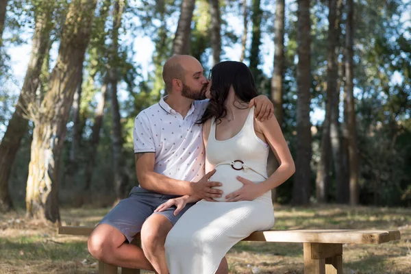 young pregnant woman, with her partner, sitting on a bench in the field.