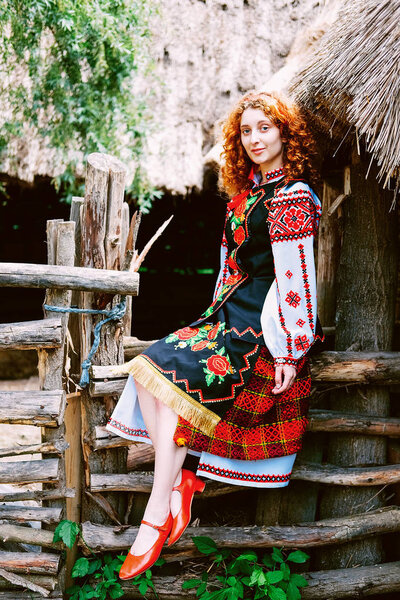 Young Slavonic woman in traditional embroidered costume and red shoes sitting on the porch