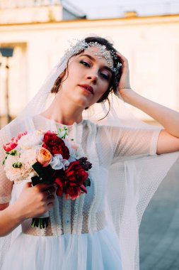 Beyaz duvak ve gelin buketi açık havada günbatımında poz tutan postiş güzel Doğu tarzı gelinle