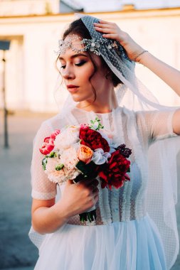 Beyaz duvak ve gelin buketi açık havada günbatımında poz tutan postiş güzel Doğu tarzı gelinle